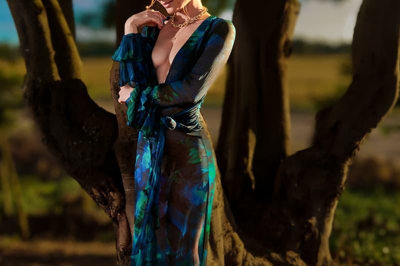Luxe escorte Bucharest in a plunging blue and green print dress with gold jewellery posing against a tree at sunset