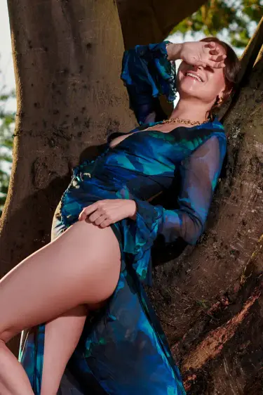 High class escort Cologne: brunette model in a blue-green print gown and gold heels reclining against a tree at sunset